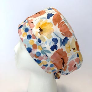 May include: A floral patterned scrub cap featuring a watercolor design with orange, yellow, and blue flowers on a white background. The cap is tied at the back and is designed to cover the head.