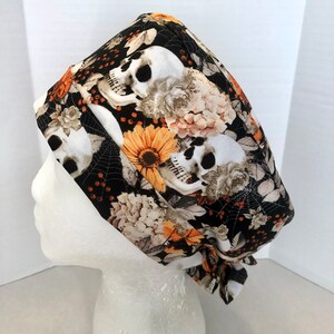 May include: Black scrub hat with a skull and floral print. The print features white skulls, orange sunflowers, and beige flowers on a black background.