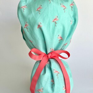 May include: A turquoise blue surgical scrub hat with a pink flamingo print and a coral pink ribbon tied around it.