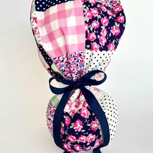 May include: A patchwork fabric scrub hat with a pink and white gingham pattern, floral print, and navy blue polka dots. The hat is tied with a navy blue ribbon.