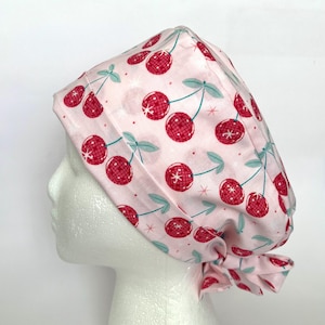 May include: A light pink surgical scrub cap featuring a pattern of red cherries with a disco ball design. The cherries have green stems and leaves, and the cap is tied at the back.