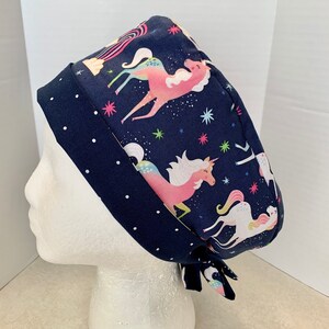 May include: A navy blue surgical scrub hat with a white polka dot trim. The hat features a colorful unicorn print with pink, blue, and yellow accents on a dark blue background.