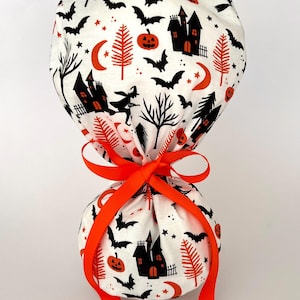 May include: A white scrub cap featuring a repeating Halloween pattern of black haunted houses, orange pumpkins, black bats, orange crescent moons, and black trees. The cap is gathered at the back and secured with a bright orange ribbon tied in a bow.