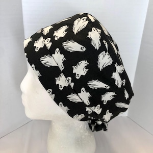 May include: A black scrub cap featuring a repeating pattern of white ghosts in various orientations. The head covering has a wide folded band at the front and a gathered back section, providing a secure fit for medical or professional use.
