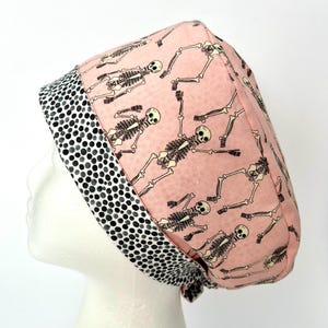 May include: A pink surgical scrub hat with a black and white polka dot trim. The hat features a repeating pattern of skeletons dancing.