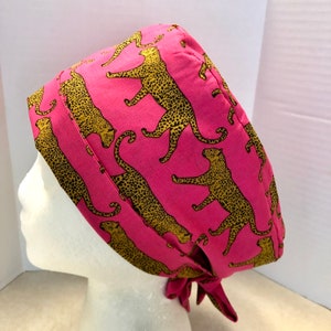 May include: A pink surgical scrub hat with a leopard print pattern. The hat has a tie at the back.