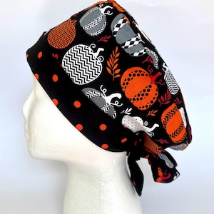 May include: A scrub cap featuring a black background with a pattern of orange, white, and gray pumpkins decorated with chevron, polka dot, and checkered designs. The wide headband is black with orange polka dots and ties at the back of the head.
