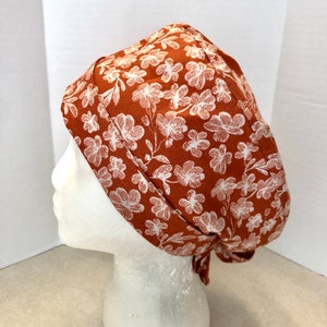 May include: A rust-colored scrub hat with a white floral pattern. The hat has a tie at the back.