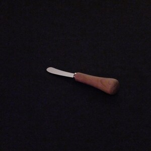 May include: A butter knife featuring a stainless steel blade and a wooden handle. The handle is a warm brown hue and has a rounded form. The blade is curved and has a smooth, shiny surface. Designed for spreading butter or soft spreads.