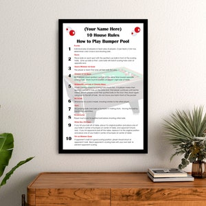 May include: A black framed poster with white text outlining 10 house rules for playing bumper pool. The poster has a red and white circle with a red dot in the center at the top. The poster is hanging on a wall above a wooden table.