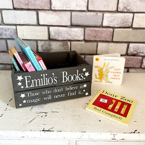 May include: A dark gray wooden crate labeled "Emilio's Books" with white text and star accents, holding several children's books. Two small books are next to the crate. The crate has the quote "Those who don't believe in magic will never find it..."