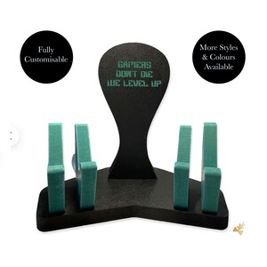 May include: Black wooden stand with four teal-coloured slots for holding controllers. The stand has a black sign with the text "Gamers Don't Die We Level Up" in green.