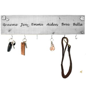 May include: A white wooden key rack with silver hooks. The names "Graeme Jen Emma Aiden Bree Bella" are printed on the rack. Two hearts and a paw print are on the rack. Keys and a dog lead hang from the hooks.