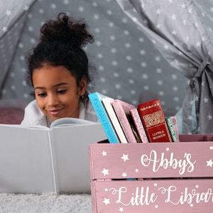 May include: A young girl reads a book in a cozy reading nook. A pink wooden crate labeled "Abby's Little Library" holds several books, including "Fairytales for Girls" and "Little Women". The crate is decorated with white stars.