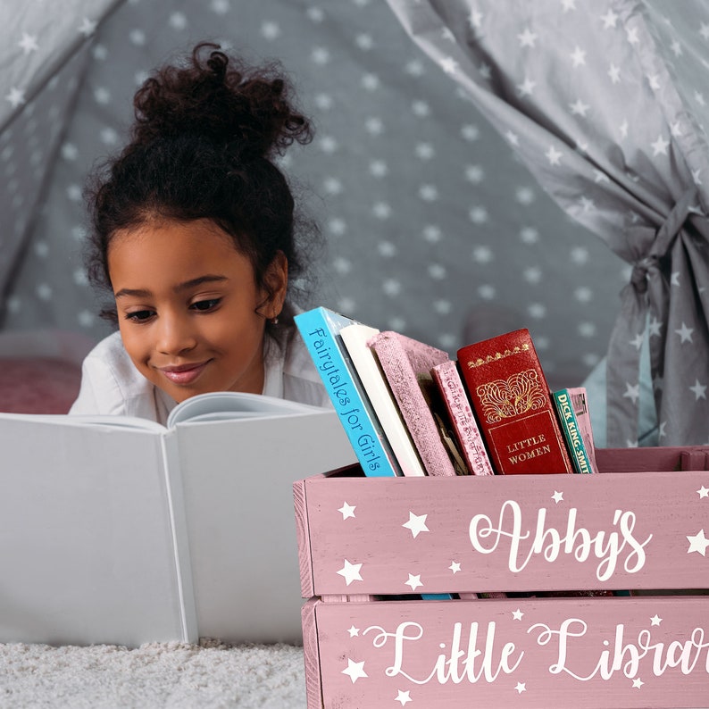 May include: A young girl reads a book in a cozy reading nook. A pink wooden crate labeled "Abby's Little Library" holds several books, including "Fairytales for Girls" and "Little Women". The crate is decorated with white stars.