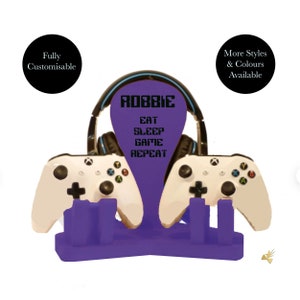 May include: Purple gaming console stand with two white controllers and a headset holder. The stand has the name "ROBBIE" and the text "EAT SLEEP GAME REPEAT" printed on it.