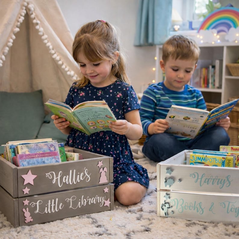May include: Two children reading books near wooden crates labeled "Hallie's Little Library" and "Harry's Books & Toys." The crates are filled with books and decorated with stars, bunnies, and angel figures. A rainbow decoration is visible in the background.