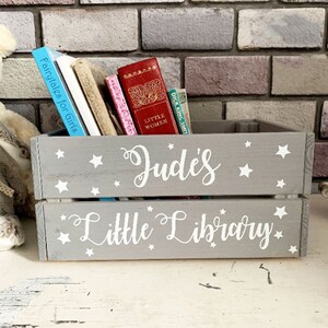 May include: A gray wooden crate labeled "Jude's Little Library" with white script and star accents. The crate holds several books, including "Fairytales for Girls" and "Little Women". The crate is painted gray with white stars.