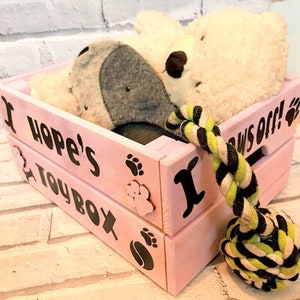 Puede incluir: Una caja de juguetes de madera rosa con las palabras "Hope's Toy Box" y huellas de patas. La caja contiene un perro de peluche y un oso de peluche blanco. Un juguete de cuerda con rayas negras, amarillas y blancas est&aacute; delante de la caja.