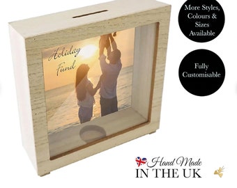 May include: White wooden photo frame with a glass front and a slot on the top for coins. The frame has a photo of a family of three on a beach with the text "Holiday Fund" written on the glass.