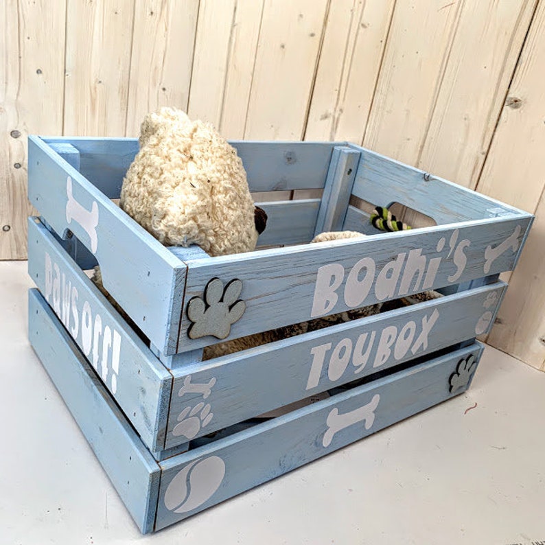 Puede incluir: Una caja de madera azul claro con texto blanco y decoraciones tem&aacute;ticas de perros. La caja dice "Bodhi's TOYBOX" y "PAWS OFF!". Presenta huellas de patas, huesos y un dise&ntilde;o de pelota de tenis. Dentro hay una cama y un juguete para perros esponjosos.