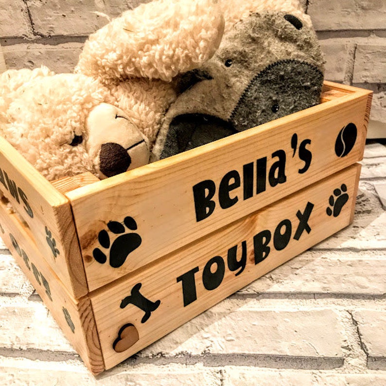 Puede incluir: Un ba&uacute;l de juguetes de madera con las palabras "Bella's TOYBOX" en letras negras. La caja est&aacute; llena de dos peluches. Huellas de patas y un coraz&oacute;n tambi&eacute;n est&aacute;n en la caja.