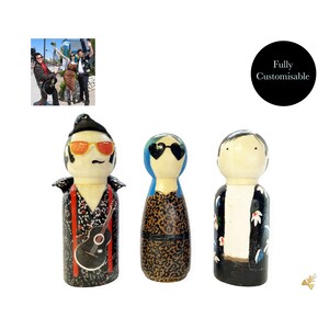 May include: Three hand-painted wooden peg dolls, each with unique clothing and accessories. The doll on the left is wearing a black and white patterned shirt with a red scarf, sunglasses, and a guitar. The middle doll is wearing a blue headscarf, sunglasses, and a leopard print dress. The doll on the right is wearing a black and white patterned shirt with white flowers, sunglasses, and black trousers.