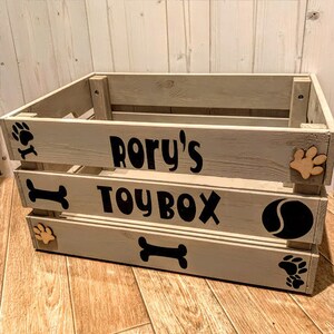 Puede incluir: Una caja de juguetes de madera gris claro con el texto "RORY'S TOYBOX" en negro. La caja est&aacute; decorada con huellas de patas, un hueso y una pelota. La caja de juguetes est&aacute; dise&ntilde;ada para juguetes de mascotas.