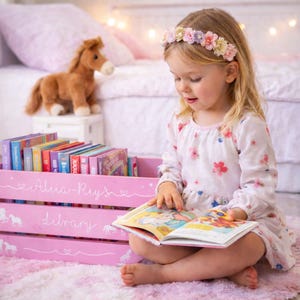 May include: A young child wearing a floral headband and dress sits on a pink rug, reading a book. A pink wooden crate labeled "Alia-Ruy's Library" holds colorful books. A stuffed horse toy sits nearby, adding to the whimsical scene.