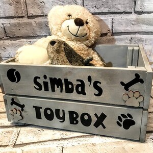 Puede incluir: Una caja de juguetes de madera gris con el nombre "Simba's" y "TOY BOX" en letras negras. La caja est&aacute; decorada con un hueso, huellas de patas y una pelota de tenis. Un oso de peluche y un b&uacute;ho est&aacute;n dentro.