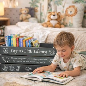 May include: A child's reading nook featuring a gray wooden crate labeled "Logan's Little Library" filled with colorful books. The crate has decorative animal illustrations and text. A child is reading a book on the floor.