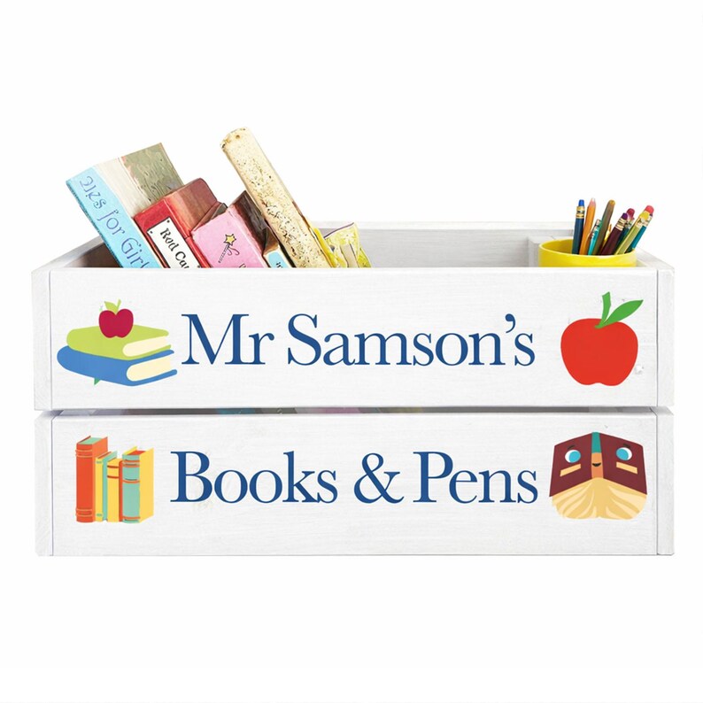 May include: White wooden storage crate with the text "Mr Samson's" and "Books & Pens" in blue. The top section contains books and a yellow cup with pencils. The crate is decorated with apple and book graphics.