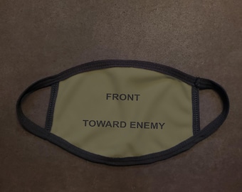 Może przedstawiać: Zielona maska ​​na twarz z czarnymi gumkami i nadrukiem "FRONT" i "TOWARD ENEMY" z przodu.