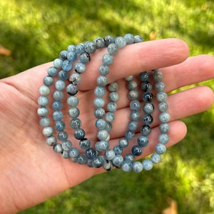 May include: Several beaded bracelets with a light blue and gray color scheme are held in a hand. The bracelets are made of small, round beads. The background is a blurred view of green grass.