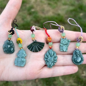 May include: Six small green stone charms featuring carved designs of a fan, a cat, a fox, and figures, each attached to a colorful knotted cord with a small green bead. The charms are dark green and translucent, displaying intricate carvings.
