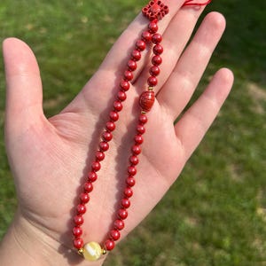 May include: A decorative lanyard featuring a loop of small, round, red beads accented with tiny gold-toned spacers. The strand includes a larger, textured, oval-shaped red bead, a single smooth, pale yellow bead at the base, and a red knotted cord top.