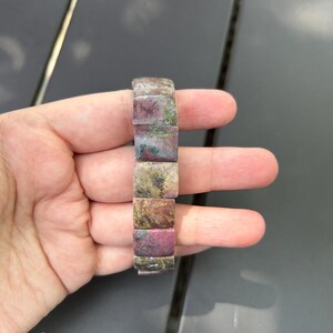 May include: A rectangular bracelet composed of multiple stone segments. The bracelet features a mix of colors, including pink, green, and brown. The bracelet is held in a hand.