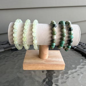 May include: Three beaded bracelets on a wooden display stand. Two bracelets are light green and one bracelet is green with black and white accents.