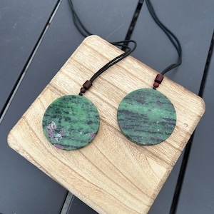 May include: Two round green and black gemstone pendants with red accents on black cords. The pendants are hanging from a wooden surface.