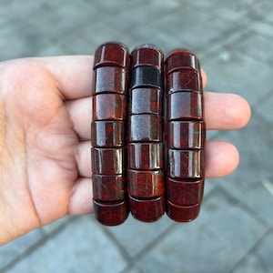 May include: Three beaded bracelets with a dark red and black color scheme. Each bracelet is made of cylindrical beads. The bracelets are held in a hand, showcasing the rich color and texture of the jewelry.