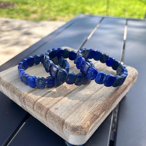 May include: Three blue lapis lazuli bracelets stacked on a wooden surface. The bracelets are made of round beads.