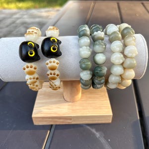 May include: Several beaded bracelets displayed on a light gray jewelry stand. The bracelets feature cat and paw-shaped beads in black, cream, and green hues. The stand is made of wood and is set on a dark surface.
