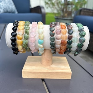 May include: A display of heart-shaped stone bracelets in various colors, including black, yellow, green, pink, and orange. The bracelets are arranged on a white fabric-covered stand with a wooden base, set on a gray table. The background includes outdoor seating.