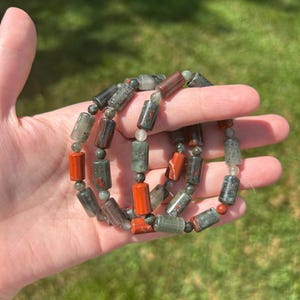 May include: A collection of beaded bracelets featuring a mix of cylindrical and round beads. The bracelets showcase a color palette of red, gray, and green. The beads are strung together, creating a layered effect.