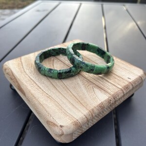 May include: Two green gemstone bracelets with a mottled pattern, stacked on a wooden surface.