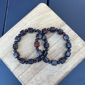 May include: Two beaded bracelets. Each bracelet features dark red and black square and round beads. The bracelets are displayed on a light-colored wooden surface against a dark blue background.