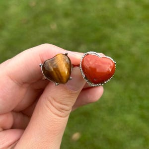 May include: Two heart-shaped rings featuring polished gemstones. One ring displays a brown and gold tiger's eye stone with a chatoyant luster, while the other features a smooth, solid red stone set within a silver-toned metal frame with a beaded edge.