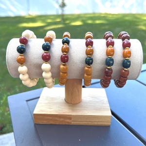 May include: Six beaded bracelets displayed on a light beige cylindrical stand. The bracelets feature a variety of colors, including cream, red, dark green, and brown, with gold-colored accents. The stand is supported by a wooden base.