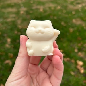 May include: A small, off-white figurine of a Maneki-neko cat, a traditional Japanese lucky charm. The cat is in a seated position with one paw raised in a beckoning gesture. The figurine has a smiling face and is held in a hand against a blurred green background.