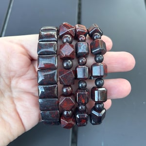 May include: Four beaded bracelets in shades of red and black. The bracelets feature various shapes, including rectangular and geometric beads. The bracelets are held in a hand against a dark background. The beads have a speckled appearance.